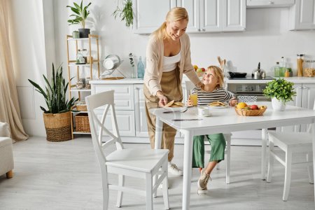 Téléchargez les photos : Mère heureuse servant des crêpes à la fille avec la jambe prothétique pendant le petit déjeuner dans la cuisine, inclusion - en image libre de droit