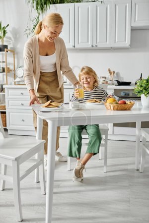Téléchargez les photos : Femme heureuse servant des crêpes à la fille avec la jambe prothétique pendant le petit déjeuner dans la cuisine, inclusion - en image libre de droit