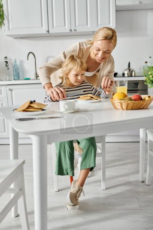 Foto de Feliz mujer de pie detrás de linda hija con la pierna protésica durante el desayuno en la cocina, inclusión - Imagen libre de derechos