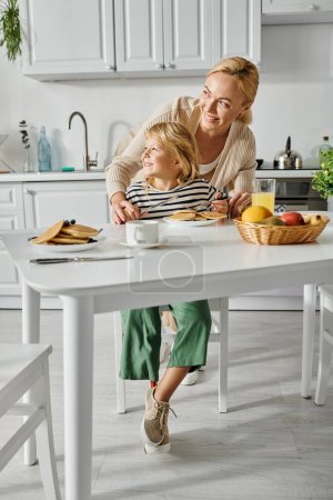 Foto de Feliz madre de pie cerca de la pequeña hija con prótesis de pierna desayunando en la cocina - Imagen libre de derechos
