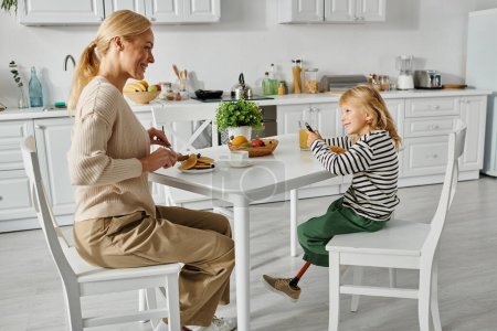 Téléchargez les photos : Mère heureuse petit déjeuner avec sa petite fille avec jambe prothétique dans la cuisine, inclusion - en image libre de droit