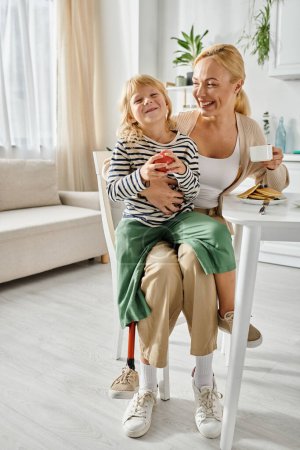 Téléchargez les photos : Fille préscolaire avec prothèse jambe assise sur les tours de mère heureuse pendant le petit déjeuner dans la cuisine - en image libre de droit