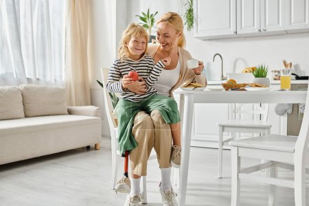 Foto de Niña en edad preescolar con la pierna protésica sentado en vueltas de madre feliz durante el desayuno en la cocina - Imagen libre de derechos