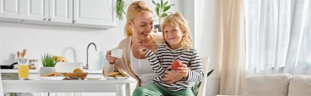 cheerful girl holding apple and sitting on laps of happy mother during breakfast in kitchen, banner
