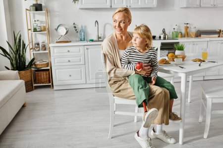 Téléchargez les photos : Fille mignonne avec la jambe prothétique assis sur les tours de mère heureuse pendant le petit déjeuner dans la cuisine - en image libre de droit