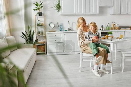 Téléchargez les photos : Petite fille avec la jambe prothétique assis sur les tours de mère blonde heureuse pendant le petit déjeuner dans la cuisine - en image libre de droit