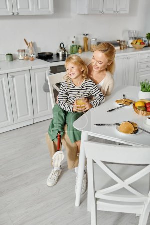 Téléchargez les photos : Préscolaire fille avec prothèse sur la jambe assis sur les tours de mère heureuse pendant le petit déjeuner dans la cuisine - en image libre de droit