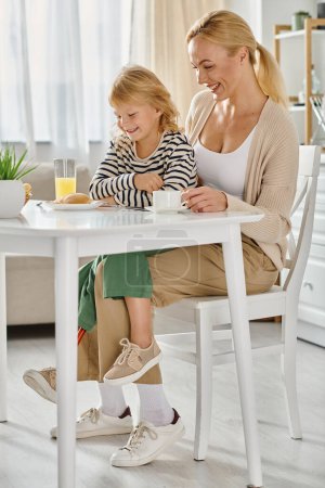 Téléchargez les photos : Enfant avec la jambe prothétique assis sur des tours de mère heureuse et regardant des crêpes pendant le petit déjeuner - en image libre de droit