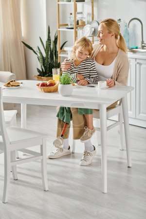 Téléchargez les photos : Fille avec la jambe prothétique assis sur des tours de mère heureuse et regardant des crêpes pendant le petit déjeuner - en image libre de droit