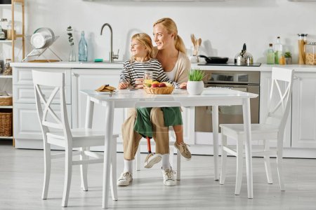 Téléchargez les photos : Préscolaire fille avec prothèse jambe assis sur les tours de mère heureuse pendant le petit déjeuner dans la cuisine - en image libre de droit
