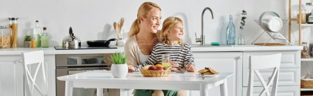 fröhliches Mädchen auf dem Schoß einer glücklichen Mutter sitzend und beim Frühstück in der Küche wegschauend, Banner