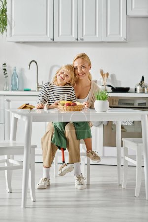 Téléchargez les photos : Mignon enfant avec la jambe prothétique assis sur les tours de mère heureuse pendant le petit déjeuner dans la cuisine - en image libre de droit