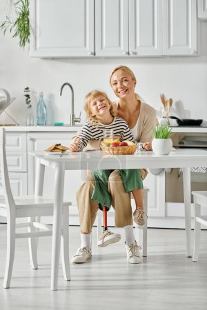 Téléchargez les photos : Fille gaie avec la jambe prothétique assis sur les tours de mère heureuse pendant le petit déjeuner dans la cuisine - en image libre de droit