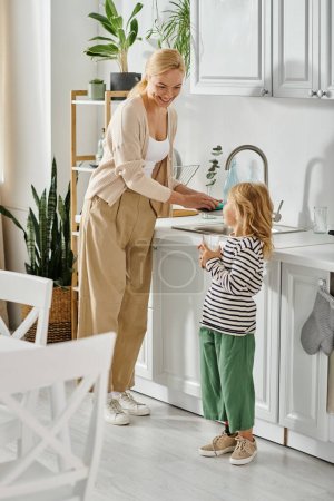 Mädchen mit Beinprothese hält ein Glas Orangensaft in der Nähe der glücklichen Mutter beim Geschirrspülen in der Küche