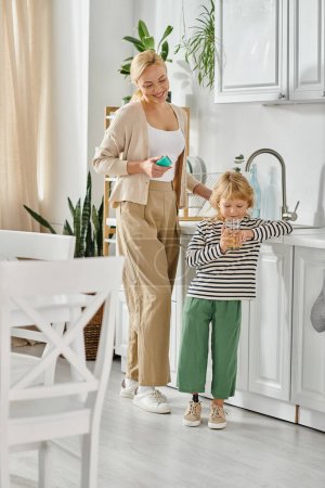 Nettes Mädchen mit Beinprothese trinkt Orangensaft in der Nähe der glücklichen Mutter beim Geschirrspülen in der Küche