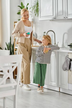girl with prosthetic leg holding glass of orange juice near happy mother washing dishes in kitchen