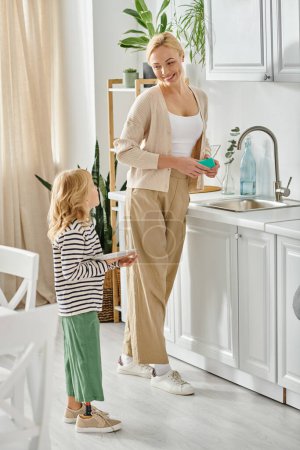 Kleines Mädchen mit Beinprothese, das Teller hält und der glücklichen Mutter beim Geschirrspülen in der Küche hilft