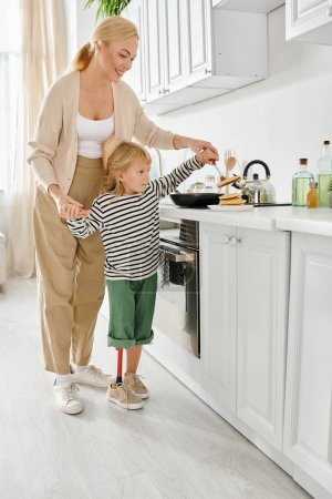 glückliche Mutter steht neben behinderter kleiner Tochter mit Beinprothese beim Pfannkuchen braten in Küche