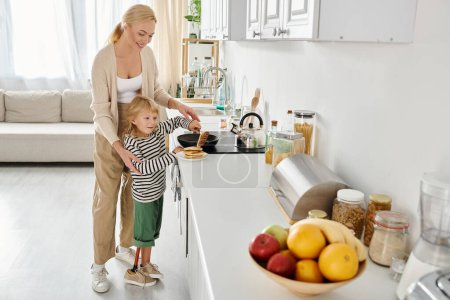 happy mother standing near little daughter with prosthetic leg frying pancakes in modern kitchen