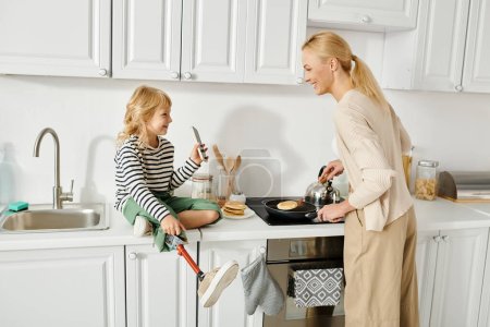 Foto de Niña con la pierna protésica sentada en la encimera y mostrando el teléfono inteligente a la madre cocinar panqueques - Imagen libre de derechos