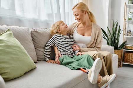 happy woman hugging daughter with prosthetic leg and sitting together on couch in living room