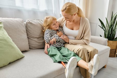 fröhliche Mutter umarmt Tochter mit Beinprothese und sitzt zusammen auf Couch im Wohnzimmer