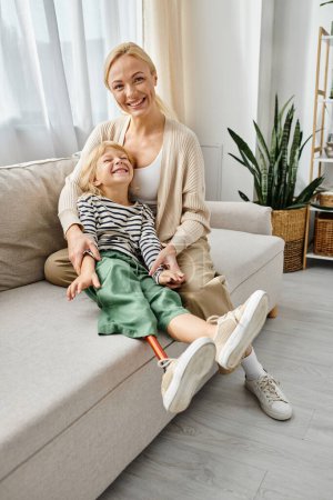 happy mother hugging cute daughter with prosthetic leg and sitting together on sofa in living room