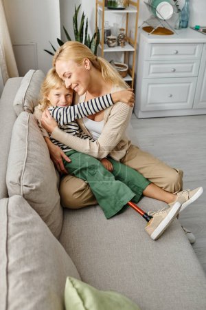 Mutter umarmt glückliche Tochter mit Beinprothese und sitzt zusammen auf Sofa im Wohnzimmer