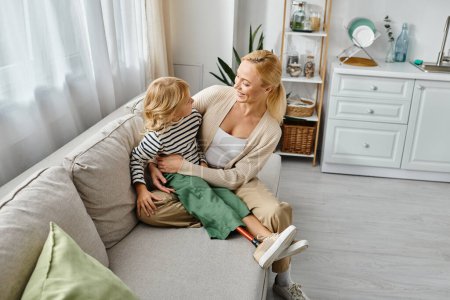freudige Mutter umarmt Tochter mit Beinprothese und sitzt zusammen auf Sofa im Wohnzimmer