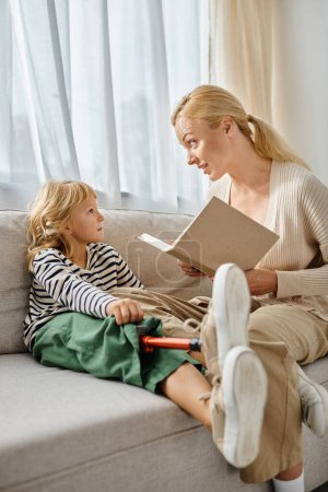 Téléchargez les photos : Blonde mère lecture livre à mignonne fille avec prothèse jambe et assis ensemble dans le salon - en image libre de droit