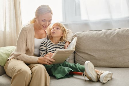 Téléchargez les photos : Mère heureuse lecture livre à sa fille avec la jambe prothétique et assis ensemble dans le salon - en image libre de droit