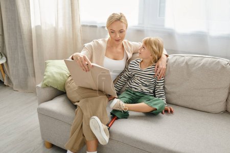 madre leyendo libro a su hija alegre con la pierna protésica y sentados juntos en la sala de estar