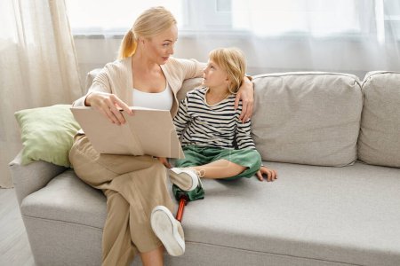 madre leyendo libro a su niño atento con la pierna protésica y sentados juntos en la sala de estar