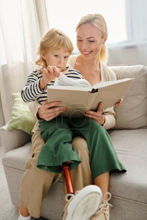 glückliches Mädchen mit Beinprothese sitzt auf dem Schoß ihrer blonden Mutter und liest Buch im Wohnzimmer