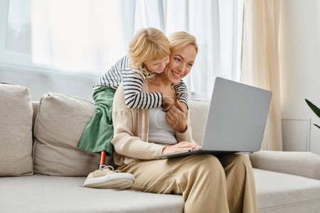 Téléchargez les photos : Enfant heureux avec prothèse jambe étreignant mère blonde travaillant sur ordinateur portable dans le salon moderne - en image libre de droit
