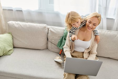 Téléchargez les photos : Enfant heureux avec prothèse jambe étreignant mère blonde travaillant sur ordinateur portable dans le salon moderne - en image libre de droit