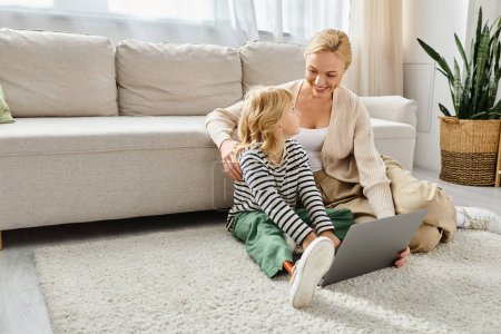 Téléchargez les photos : Enfant heureux avec la jambe prothétique à l'aide d'un ordinateur portable et assis sur le tapis avec la mère dans le salon - en image libre de droit