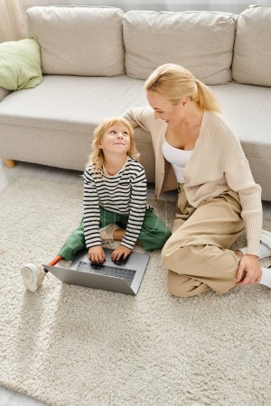 Téléchargez les photos : Enfant heureux avec la jambe prothétique à l'aide d'un ordinateur portable et assis sur le tapis avec la mère dans le salon - en image libre de droit
