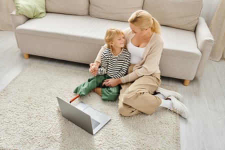 Téléchargez les photos : Fille heureuse avec prothèse jambe regarder film sur ordinateur portable et assis sur le tapis avec maman - en image libre de droit