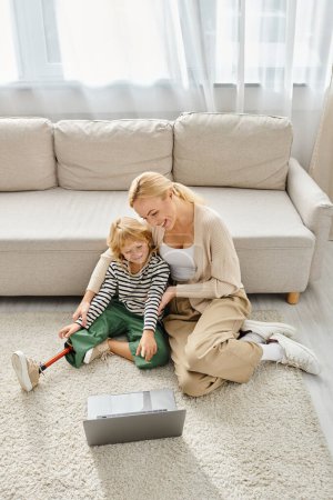 Téléchargez les photos : Enfant heureux avec prothèse jambe regarder film sur ordinateur portable et assis sur le tapis avec la mère - en image libre de droit