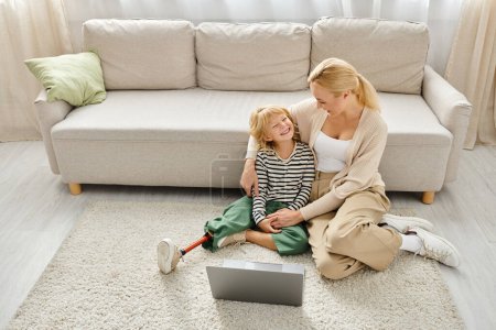 Téléchargez les photos : Fille heureuse avec prothèse jambe regarder film sur ordinateur portable et assis sur le tapis avec la mère - en image libre de droit