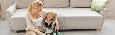 feliz mujer rubia abrazando alegre hija pequeña cerca del sofá en la sala de estar moderna, pancarta