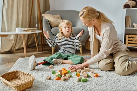 Verwirrtes Mädchen mit Beinprothese sitzt auf Teppich neben Holzspielzeug und Mutter, zuckende Geste
