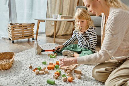 behindertes kleines Mädchen mit Beinprothese sitzt auf Teppich und betrachtet Holzspielzeug in der Nähe der Mutter