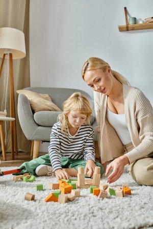 Kleines Mädchen mit Beinprothese sitzt auf Teppich und spielt mit Holzspielzeug in der Nähe der blonden Mutter
