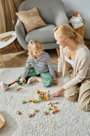 kleines Mädchen mit Beinprothese sitzt auf Teppich und spielt mit Holzspielzeug in der Nähe der glücklichen Mutter