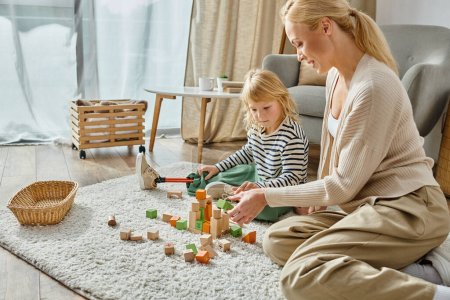 Kleines Mädchen mit Beinprothese sitzt auf Teppich und spielt mit Holzspielzeug neben freudiger Mutter