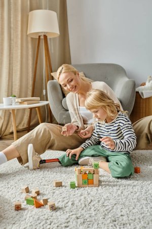 Kleines Mädchen mit Beinprothese sitzt auf Teppich und spielt mit Holzspielzeug neben fröhlicher Mutter