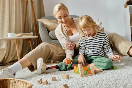 Foto de Niña preescolar con la pierna protésica sentado en la alfombra y jugando con bloques de madera cerca de la madre - Imagen libre de derechos