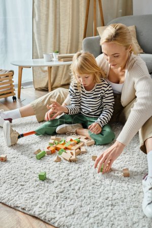 Téléchargez les photos : Fille surprise avec la jambe prothétique assis sur le tapis et jouer avec des blocs de bois près de la mère - en image libre de droit